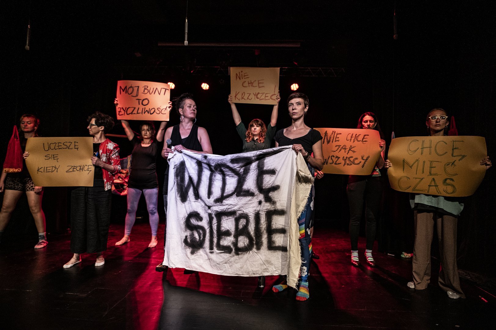 Przedstawienie teatralne. Manifestacja. Aktorki trzymają w rękach transparenty w hasłami Widzę siebie, Uczeszę się kiedy zechcę, Mój bunt to życzliwość, Nie chcę krzyczeć, Nie chcę jak wszyscy, Chcę mieć czas