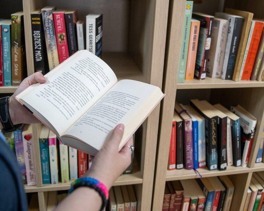 Dłonie trzymające otwartą książkę na tle regału bibliotecznego.