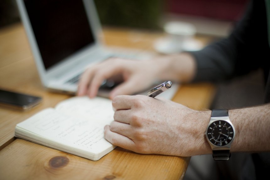 Zbliżenie na męskie dłonie trzymające długopis. Na lewej ręce czarny zegarek. Mężczyzna siedzi przy biurku i notuje coś w brulionie, w tle srebrny laptop z wyłączonym ekranem.