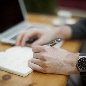 Zbliżenie na męskie dłonie trzymające długopis. Na lewej ręce czarny zegarek. Mężczyzna siedzi przy biurku i notuje coś w brulionie, w tle srebrny laptop z wyłączonym ekranem.