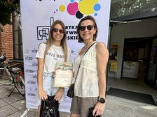 Na zdjęciu widać dwie kobiety na tle kolorowego banneru reklamowego z napisami Fabryka Aktywności Miejskiej. Z lewej strony stoi rower a z prawej widać wejście do budynku.