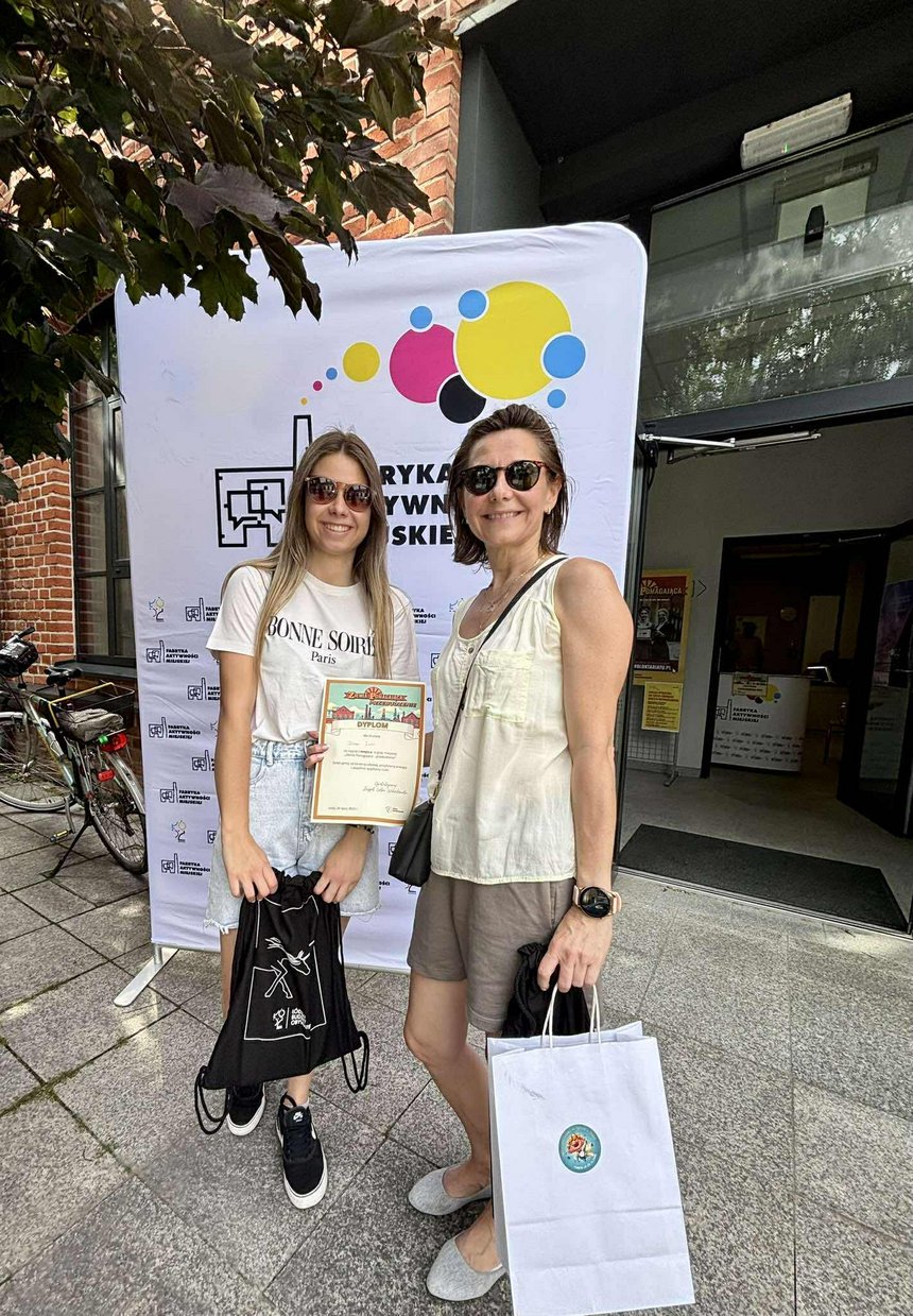 Na zdjęciu widać dwie kobiety na tle kolorowego banneru reklamowego z napisami Fabryka Aktywności Miejskiej. Z lewej strony stoi rower a z prawej widać wejście do budynku.