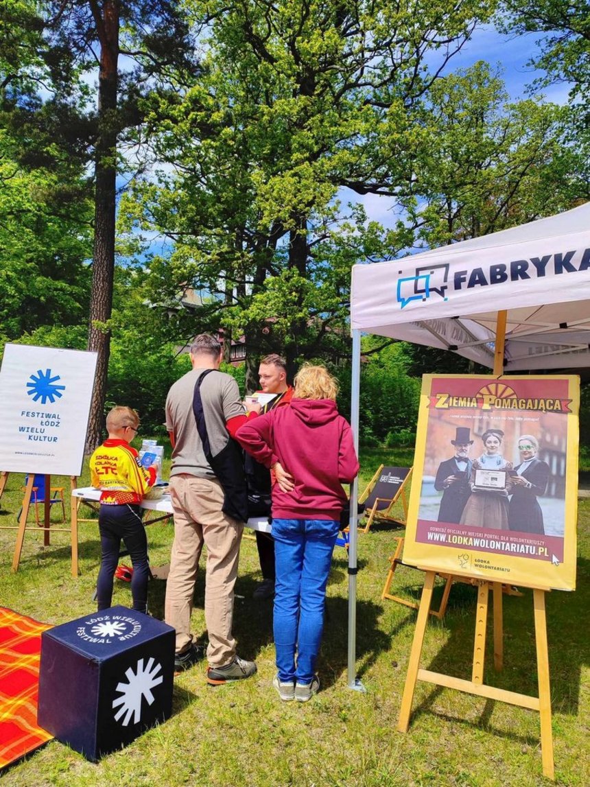 Na zdjęciu widzimy plenerowe wydarzenie odbywające się wśród zielononych drzew. Na pierwszym planie widać namiot z napisem "Fabryka", przy którym stoi kilka osób: trzy osoby dorosłe i jedno dziecko. Z lewej strony zdjęcia widać sztalugę z logo i napisem "FESTIWAL ŁÓDŹ WIELU KULTUR". Na trawie w pobliżu widać fragment koca czerwonego w żółtą kratę, na któym znajduje się sześcienny puf z takim samym napisem i logotypem jak na sztaludze. Po prawej stronie znajduje się sztaluga z plakatem, na którym widać trzy osoby w eleganckich strojach. U góry pakatu znajduje się napis "ZIEMIA POMAGAJĄCA" a u dołu adres strony internetowej www.lodkawolontariatu.pl. Pod adresem umieszczono napis ŁÓDKA WOLONTARIATU oraz logo "ŁÓDŹ kreuje"