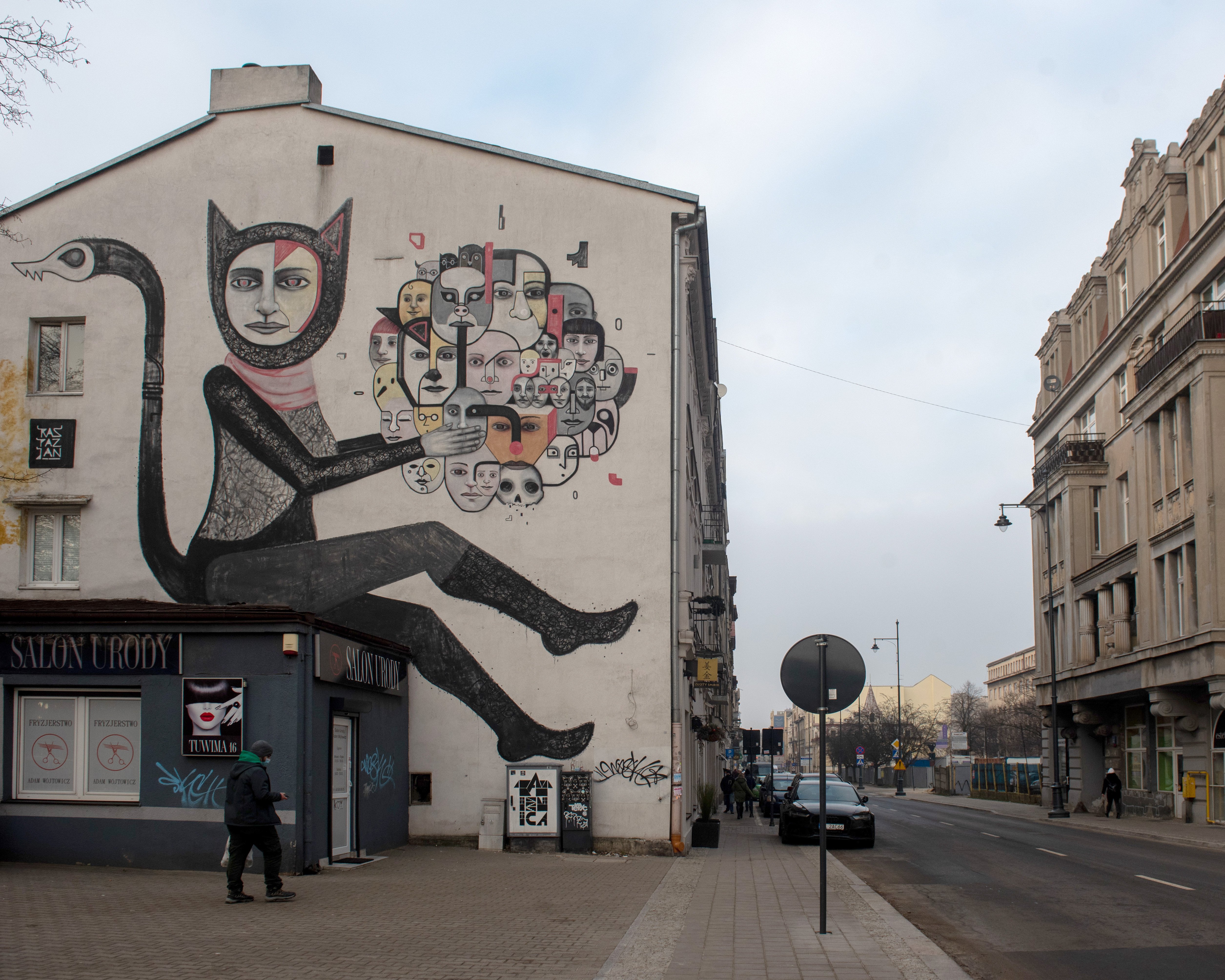  , Mural na białej ścianie budynku przedstawiający kobietę siedzącą bokiem w przebraniu kota. W dłoniach trzyma zlepek różnych twarzy.
