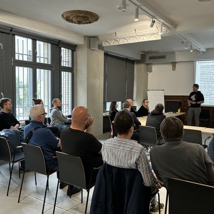 Na zdjęciu widać salę wykładową, gdzie trwa spotkanie. Na ekranie wyświetlany jest slajd z napisami. Uczestnicy siedzą na krzesłach, zwróceni w stronę ekranu. Uczestnicy spotkania siedzą na krzesłach przodem do ekranu. Przed ekranem ustawiono ławki w literę „U”, przy których siedzi kilka osób. Z lewej strony stoi osoba tyłem do ekranu, prawdopodobnie prowadzi spotkanie. Na zdjęciu widzimy piętnaście osób. Pomieszczenie jest jasne, po lewej stronie znajdują się duże okna a na suficie są dekoracje i zdobienia jak również umieszczone są punktowe światła.